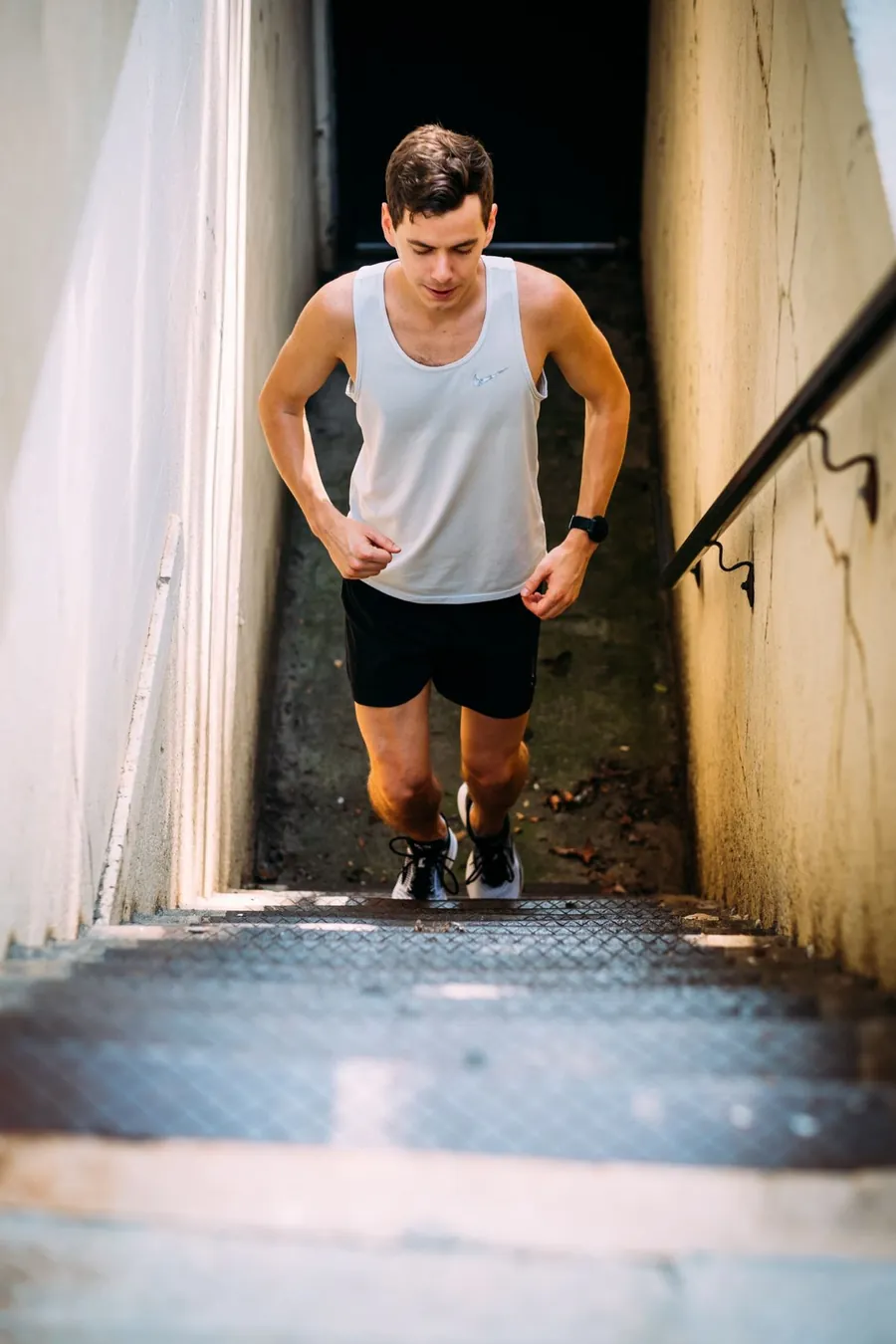 Chris running up a narrow outdoor stairwell in a white tank and black shorts